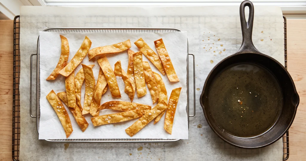 Golden crispy fried corn tortilla strips draining on a paper towel-lined tray, lightly salted, next to a cast iron skillet